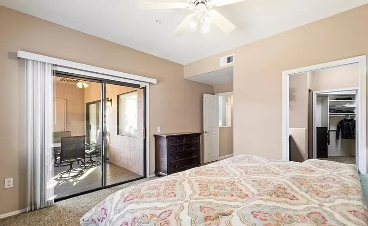 This bedroom features a bed with patterned bedding, a sliding glass door leading to a patio, and a dresser. Sunlight streams in, creating a calm, airy space ideal for morning routines or quiet evenings. The doorway hints at peaceful views and the option to enjoy fresh air.