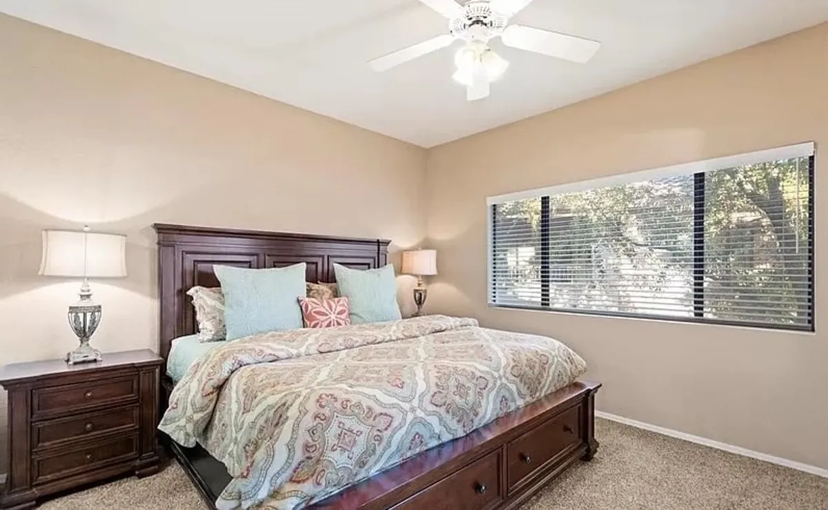 This image depicts a tranquil bedroom scene with a bed dressed in patterned bedding, flanked by decorative lamps, and a window with blinds letting in soft natural light. The atmosphere is serene and cozy, ideal for unwinding after a long day.