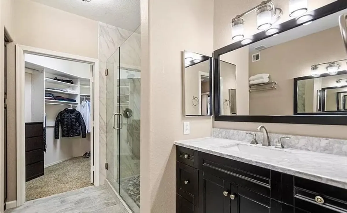 A modern bathroom with a double sink vanity, a glass shower enclosure with morning light streaming through, and a walk-in closet. The space is well-organized and refreshing, supporting personal grooming and comfort for multiple guests.